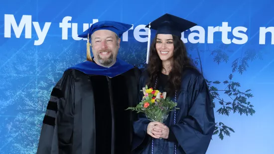 A man and woman wearing graduation regalia stand in front of a blue background.