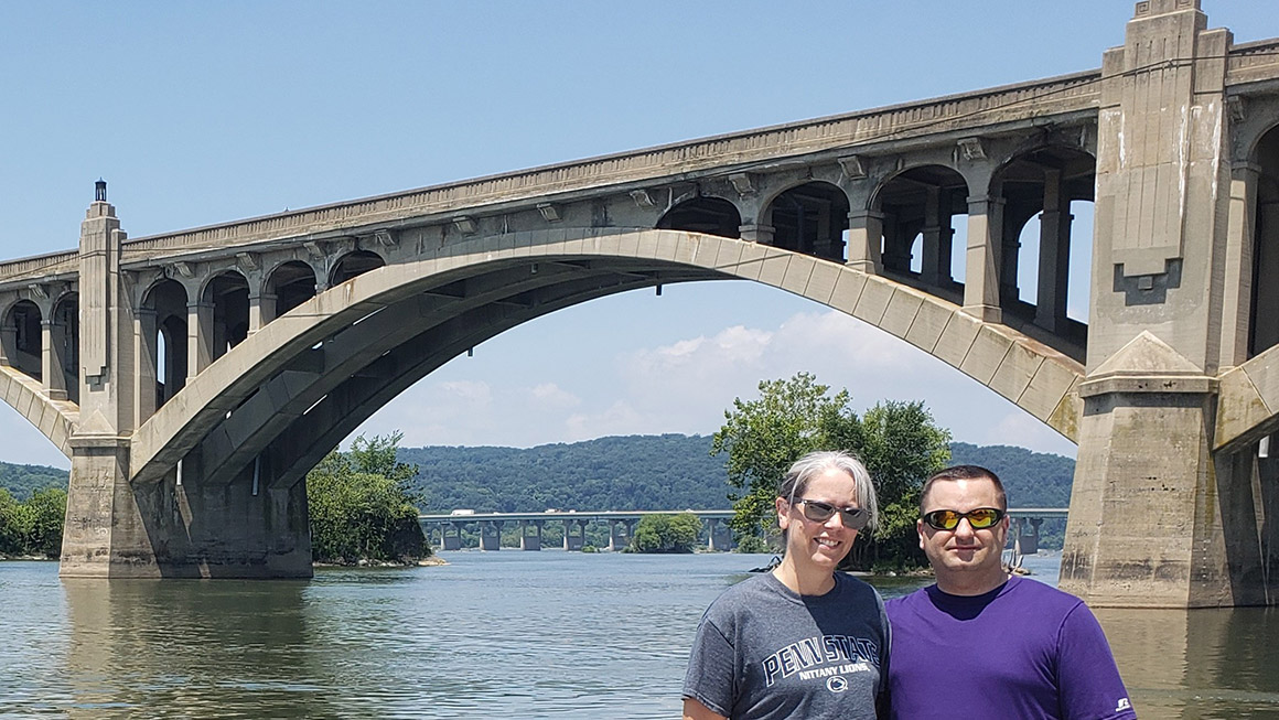 This photo shows Julie Brubaker with her husband on the banks of the Susquehanna River with a bridge in the background.