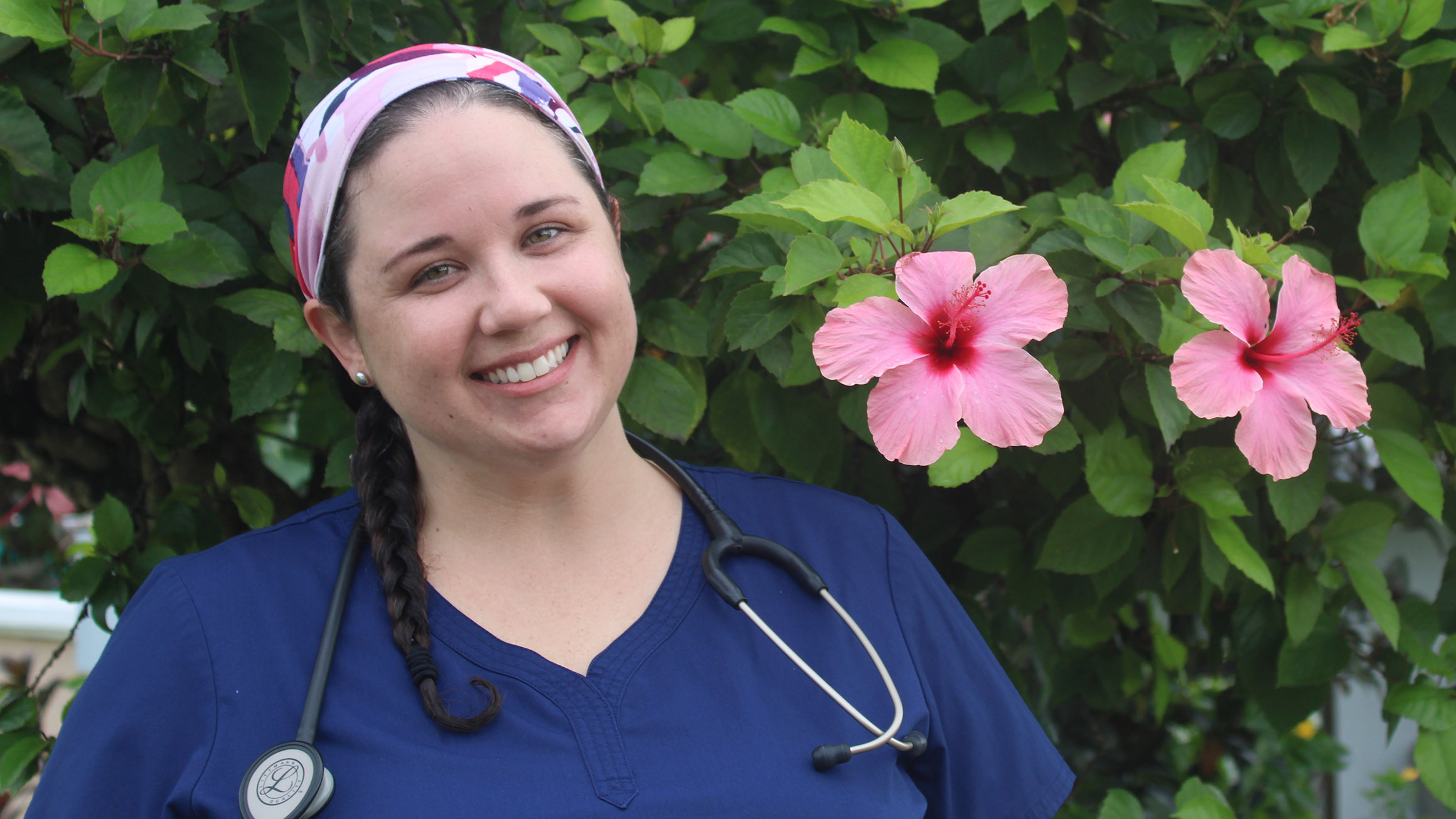 Katie Francis is wearing scrubs standing in front of a green plant with pink flowers