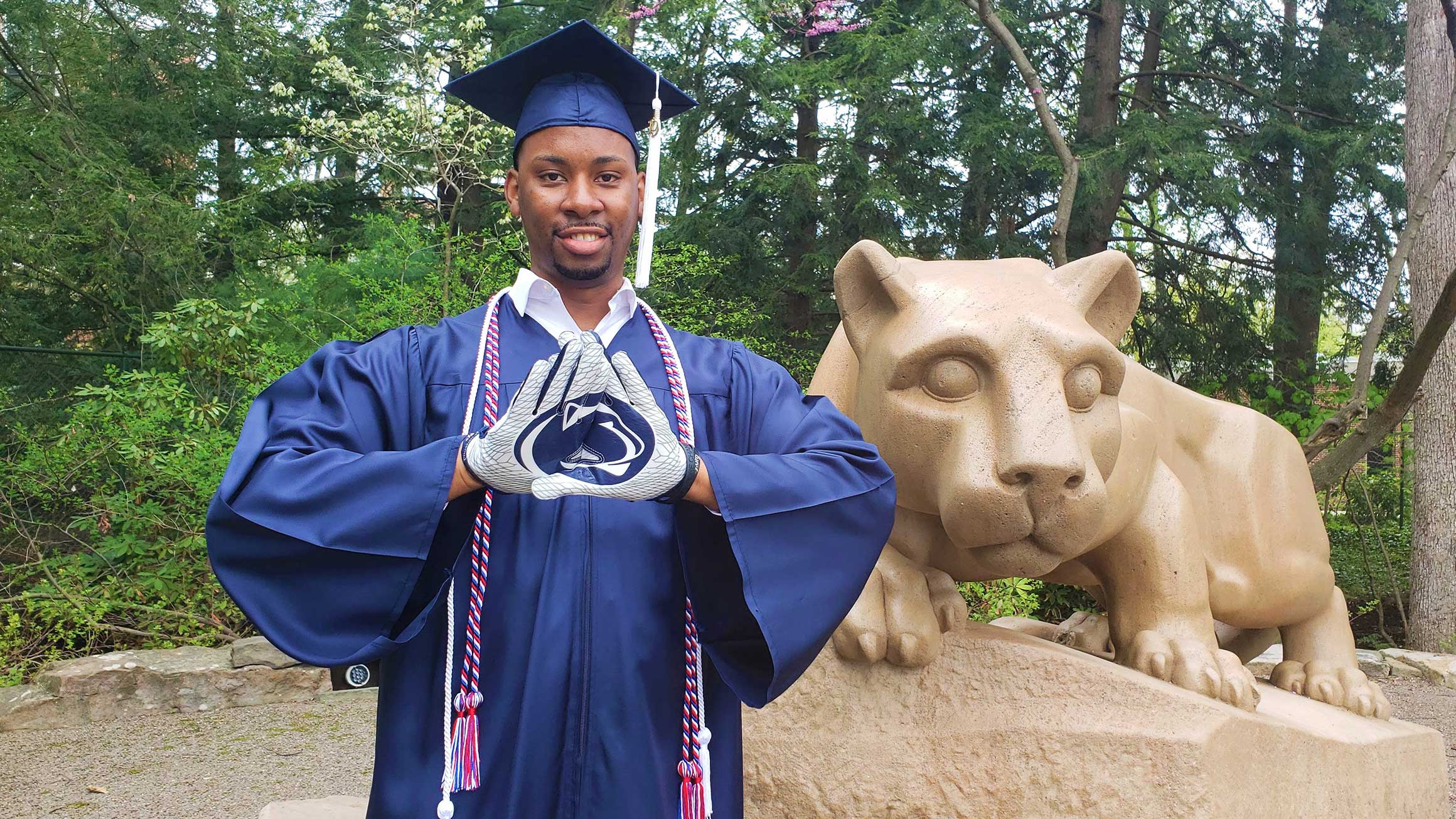 A student named James Elder wears his cap and gown in front of the Nittany Lion statue.