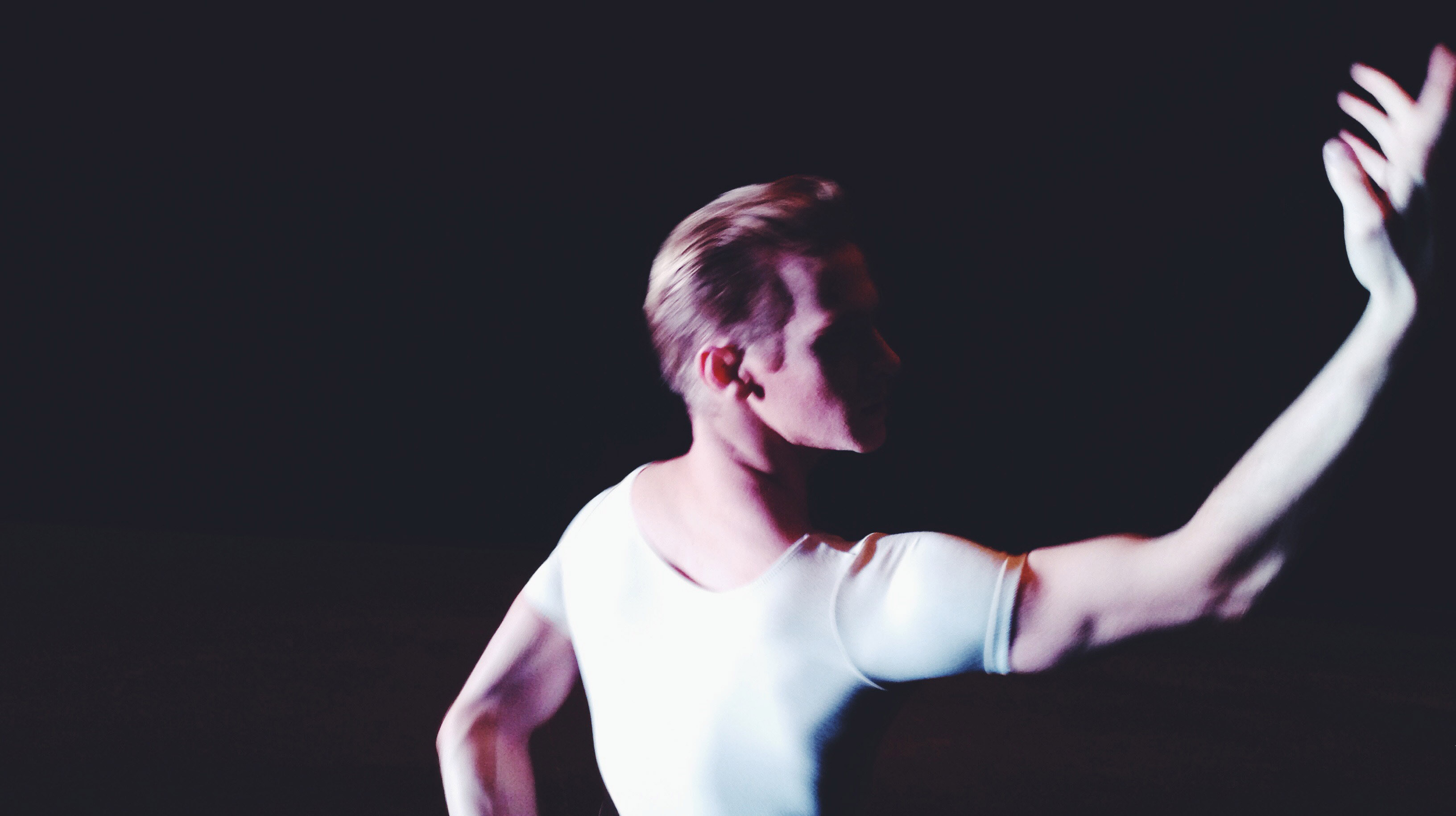 Lars Nelson doing ballet on a dark stage, with one hand raised in the air