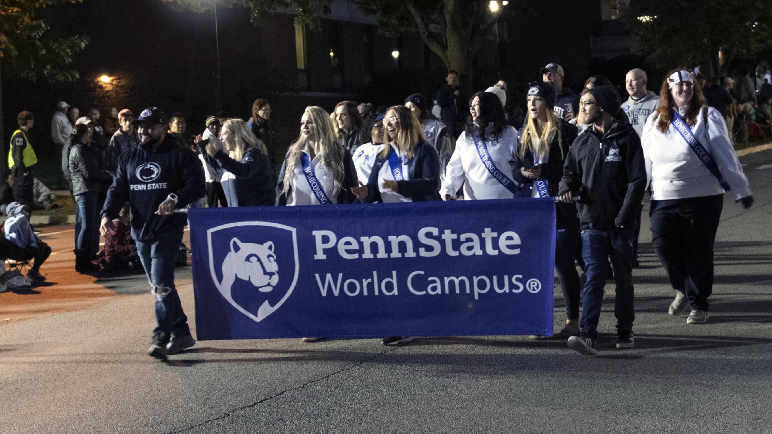 Members of the Penn State World Campus Homecoming Committee and Court walk in the homecoming parade, carrying a World Campus banner.
