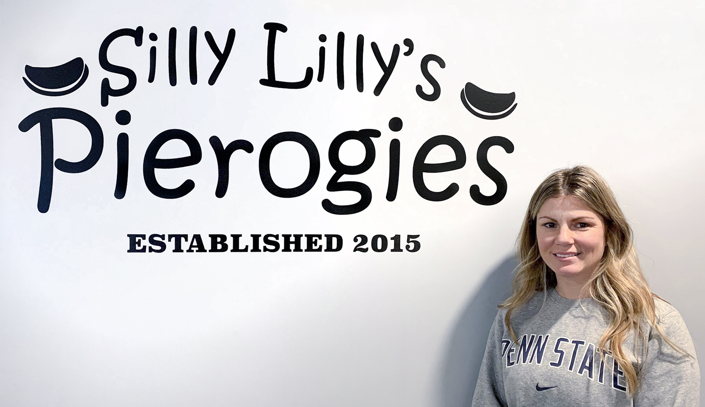 Nicole Jankowski, wearing a Penn State sweatshirt, stands beside the logo for her company, Silly Lilly's Pierogies, which is printed on a wall.