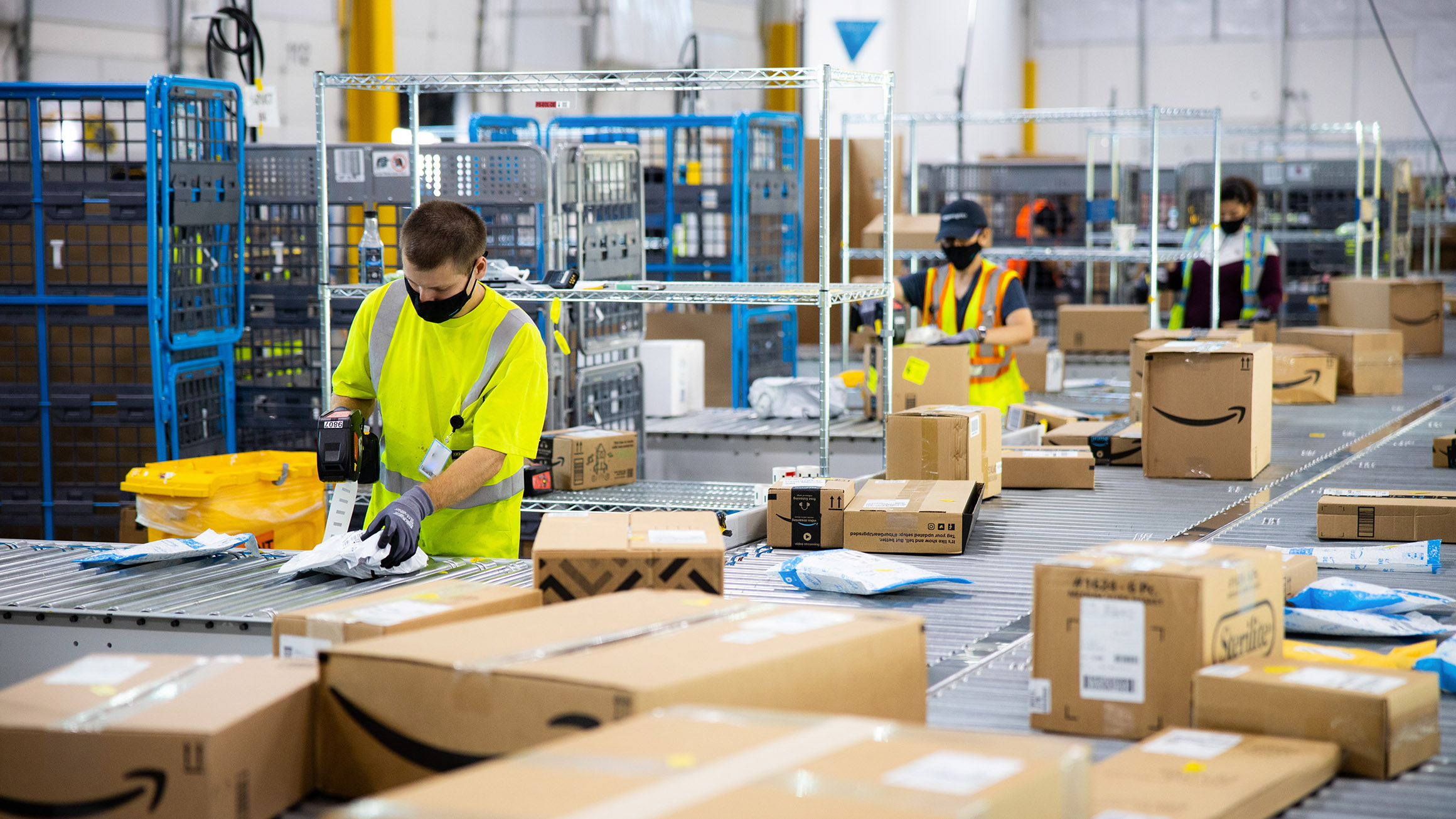 Two Amazon employees wearing masks are sorting packages in a fulfillment center.