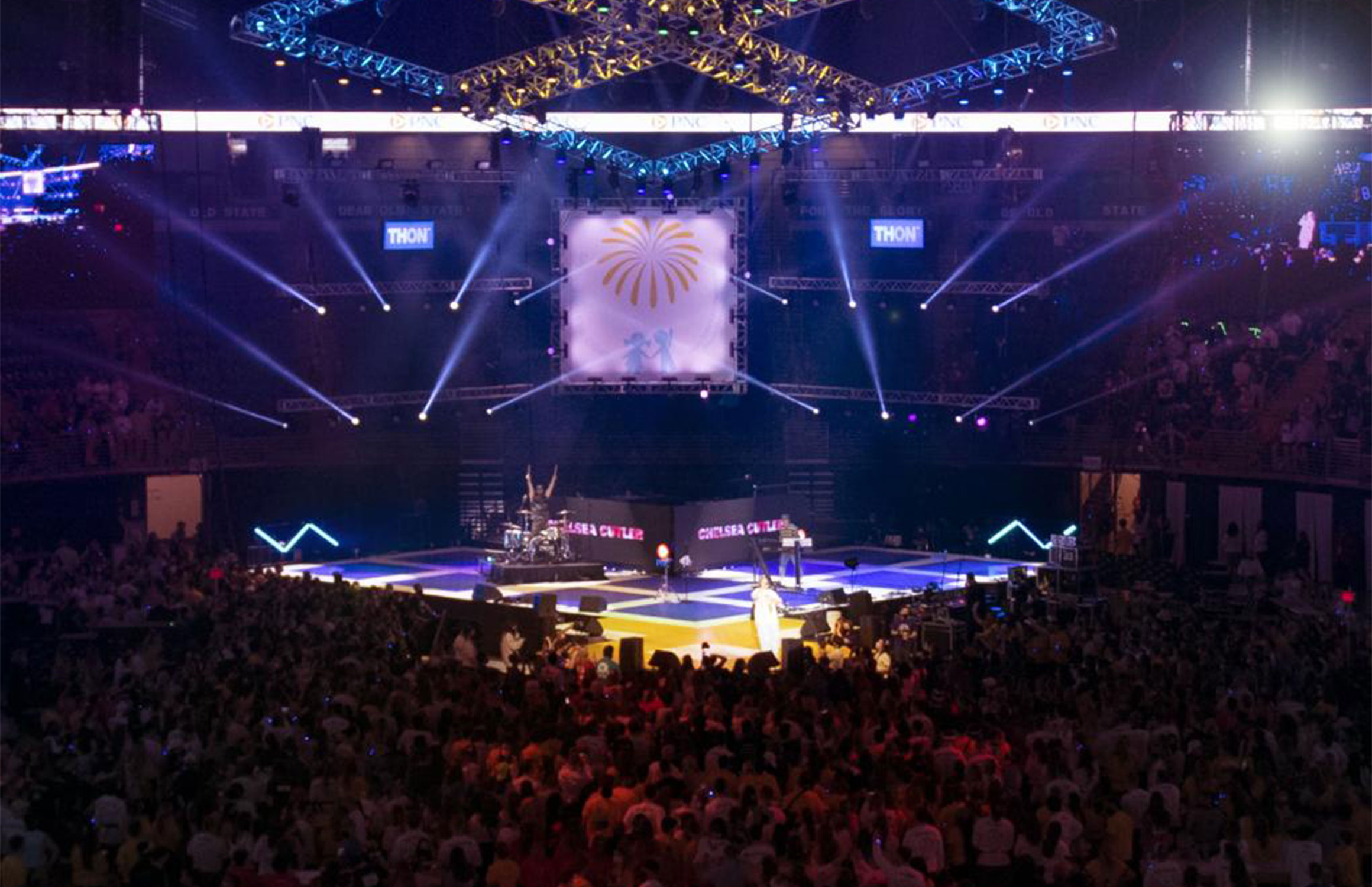 A wide shot of the dance marathon shows the stage and thousands of people seated in the Bryce Jordan Center