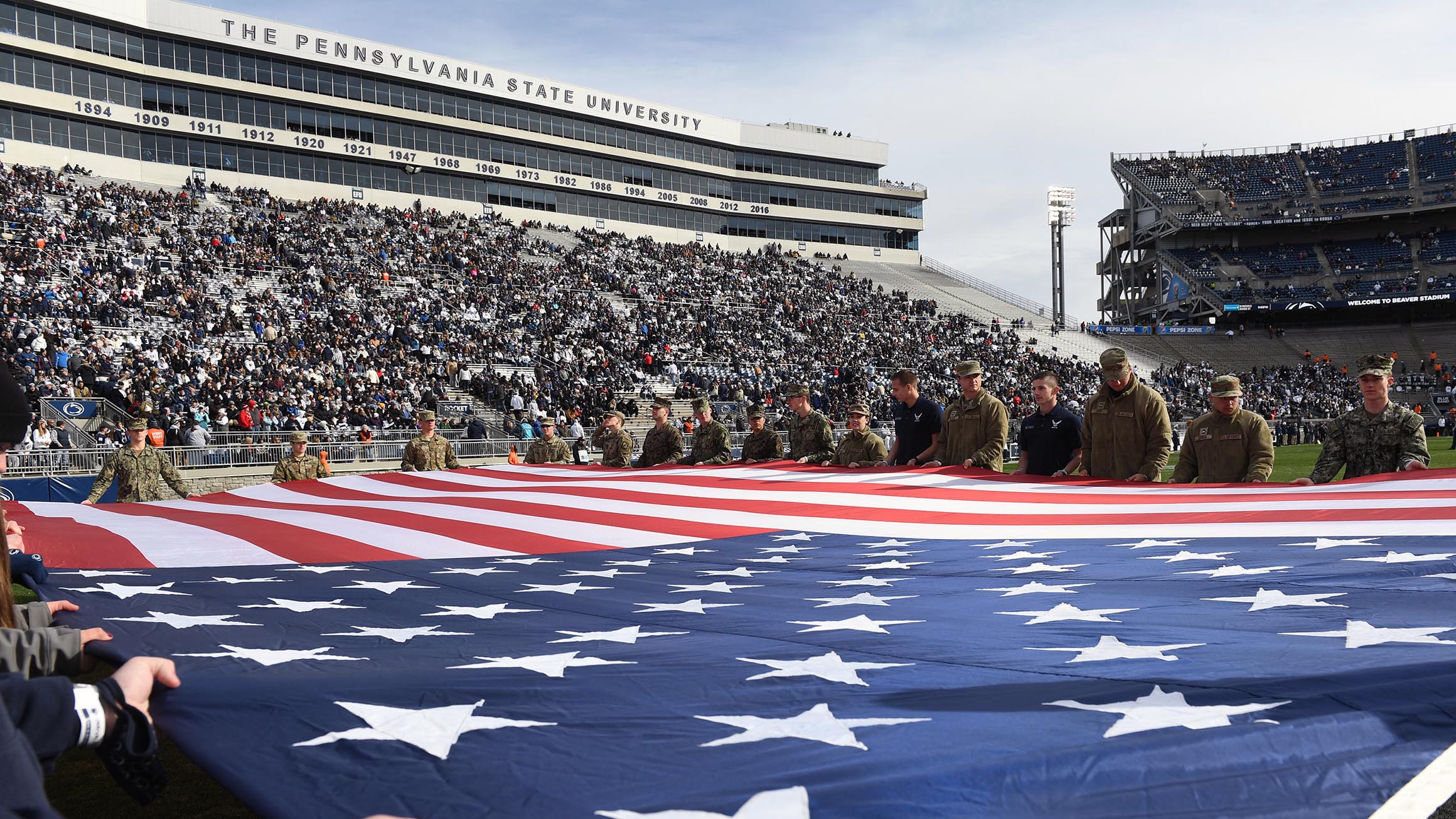 An American flag is displayed on the field at Beaver Stadium by military service members.