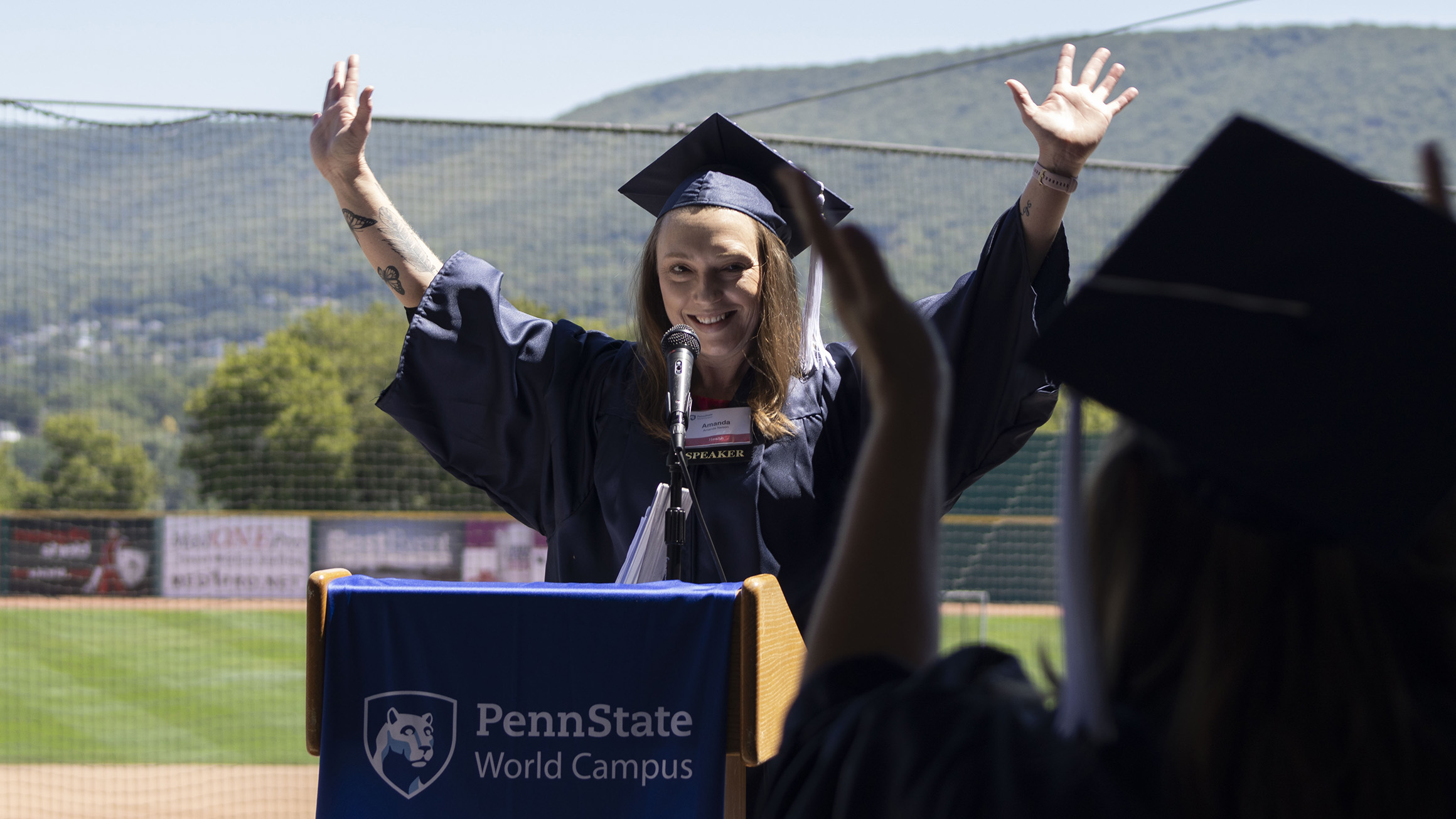 A woman wearing a blue graduation cap and gown raises her hands in celebration.
