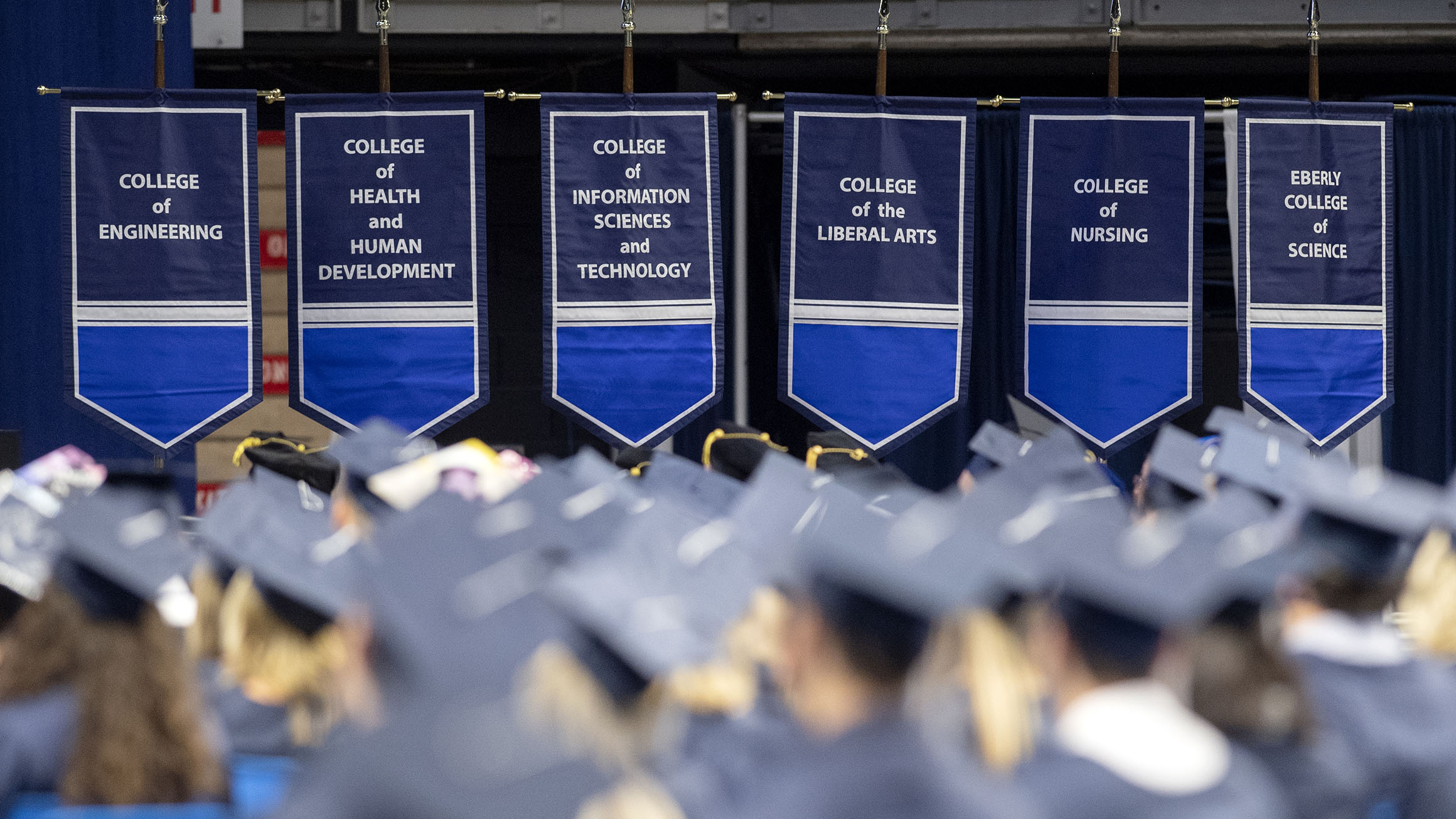 Banners with the names of colleges of Penn State hang on the graduation stage while people wearing caps and gowns are shown seated.