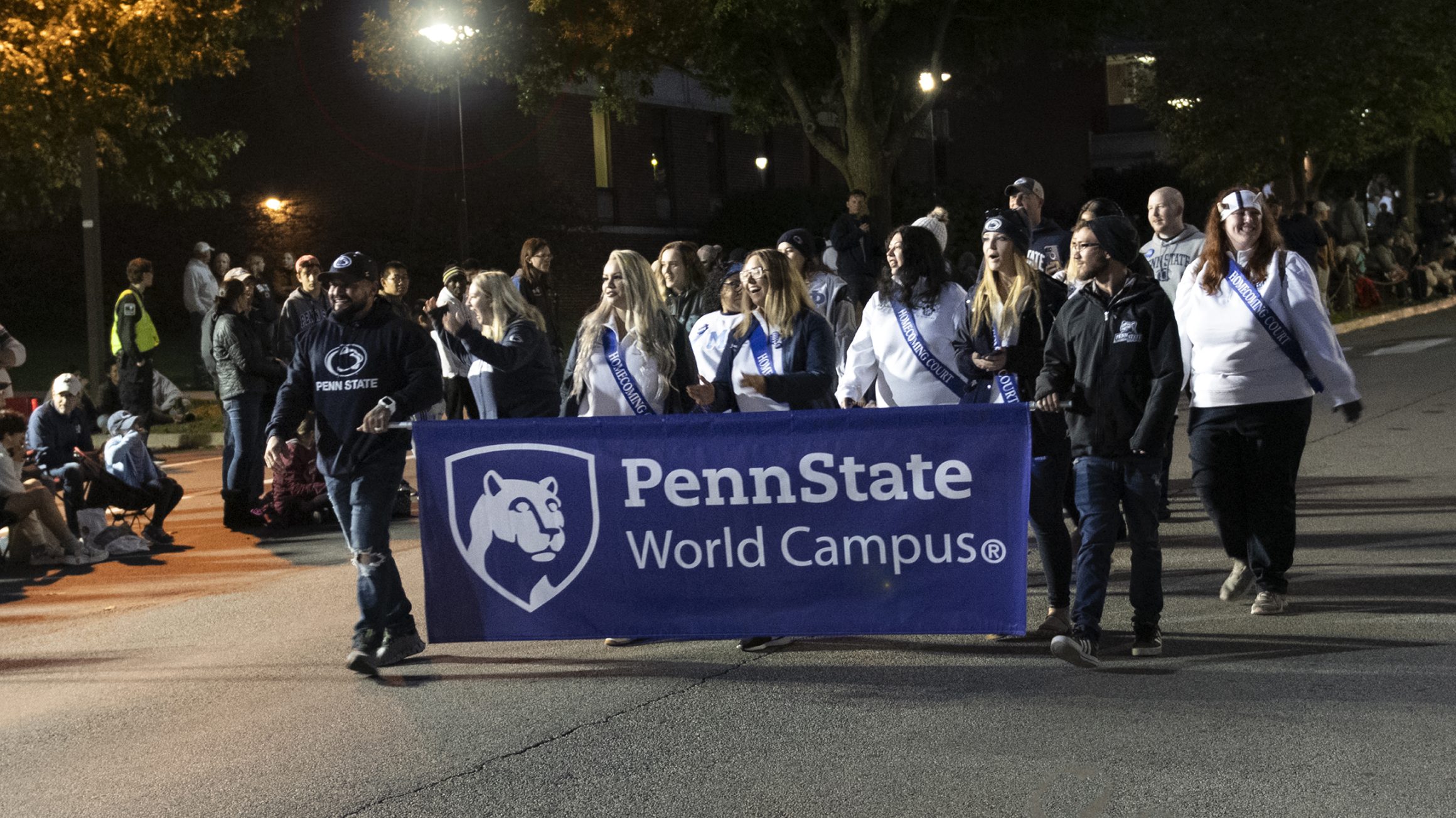 A group of people hold a blue World Campus banner in a parade at night.
