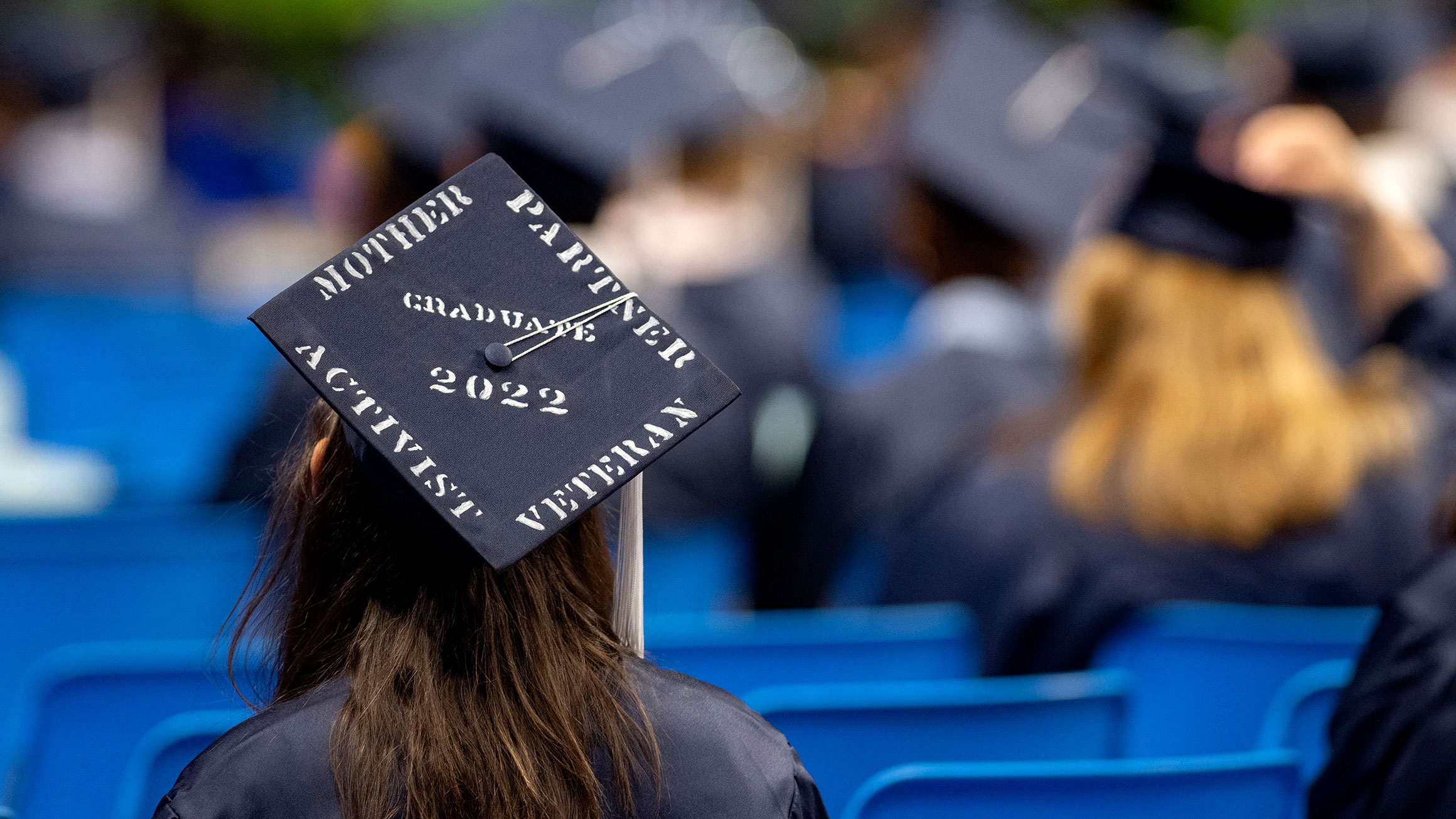 A student wears a cap that says mother, partner, activist, veteran while sitting at graduation