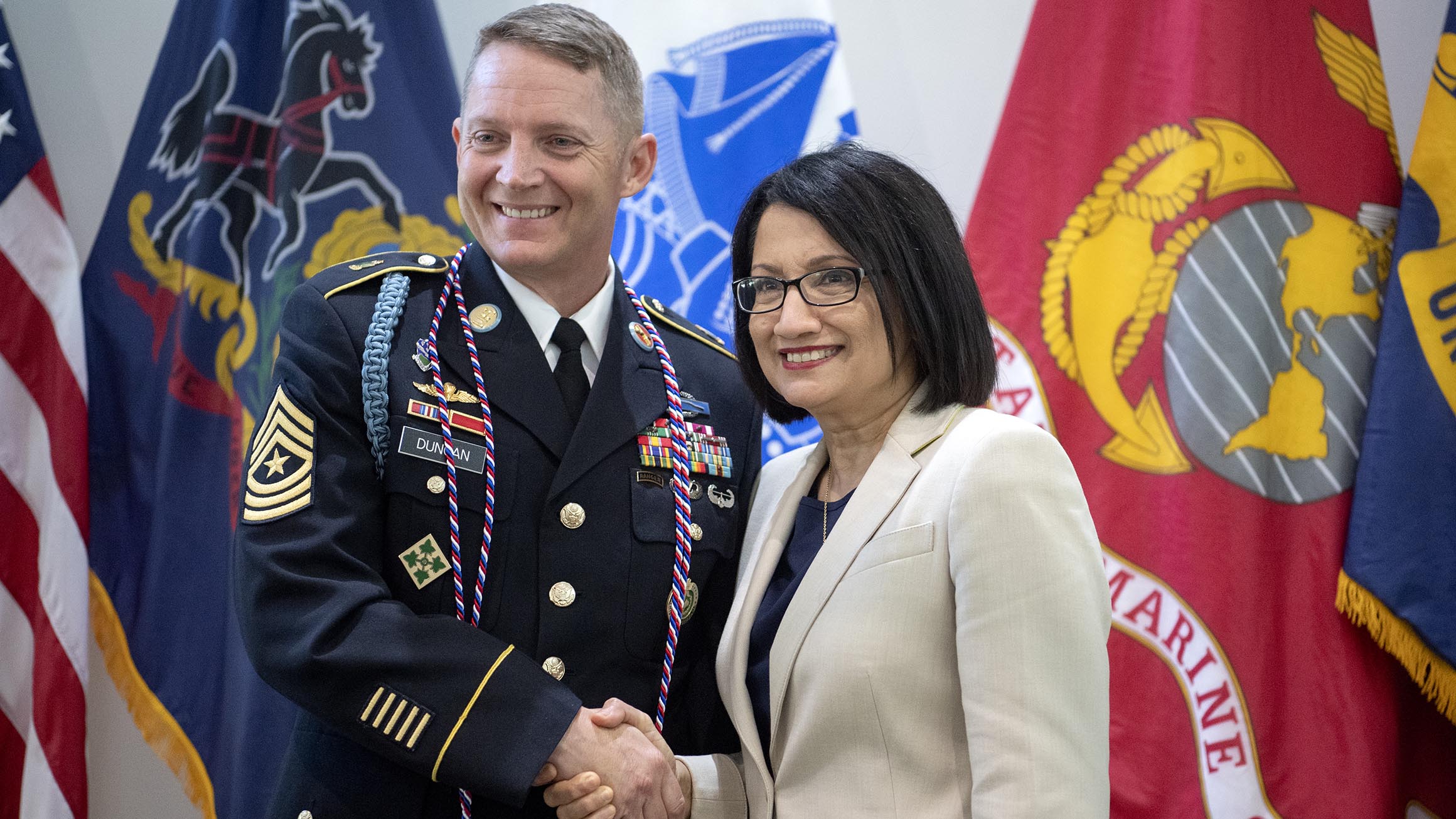 President Neeli Bendapudi shakes hands with a member of the Army with flags of the military in the background.