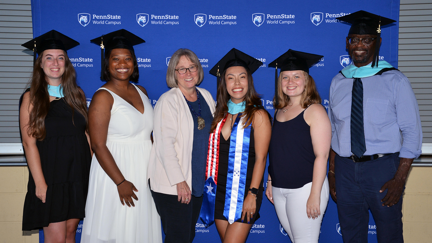 A group of five graduates and a faculty member pose in front of a blue backdrop.