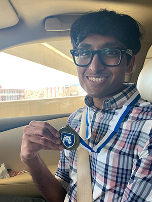 Harish Logaraj, sitting in the passenger seat of a car, holds us a medal.