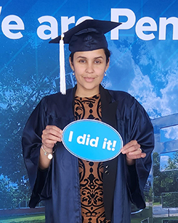 A woman wearing a blue graduation cap and gown stands in front of a blue Penn State-themed backdrop.