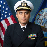 Man in Navy uniform stands in front of US flag.