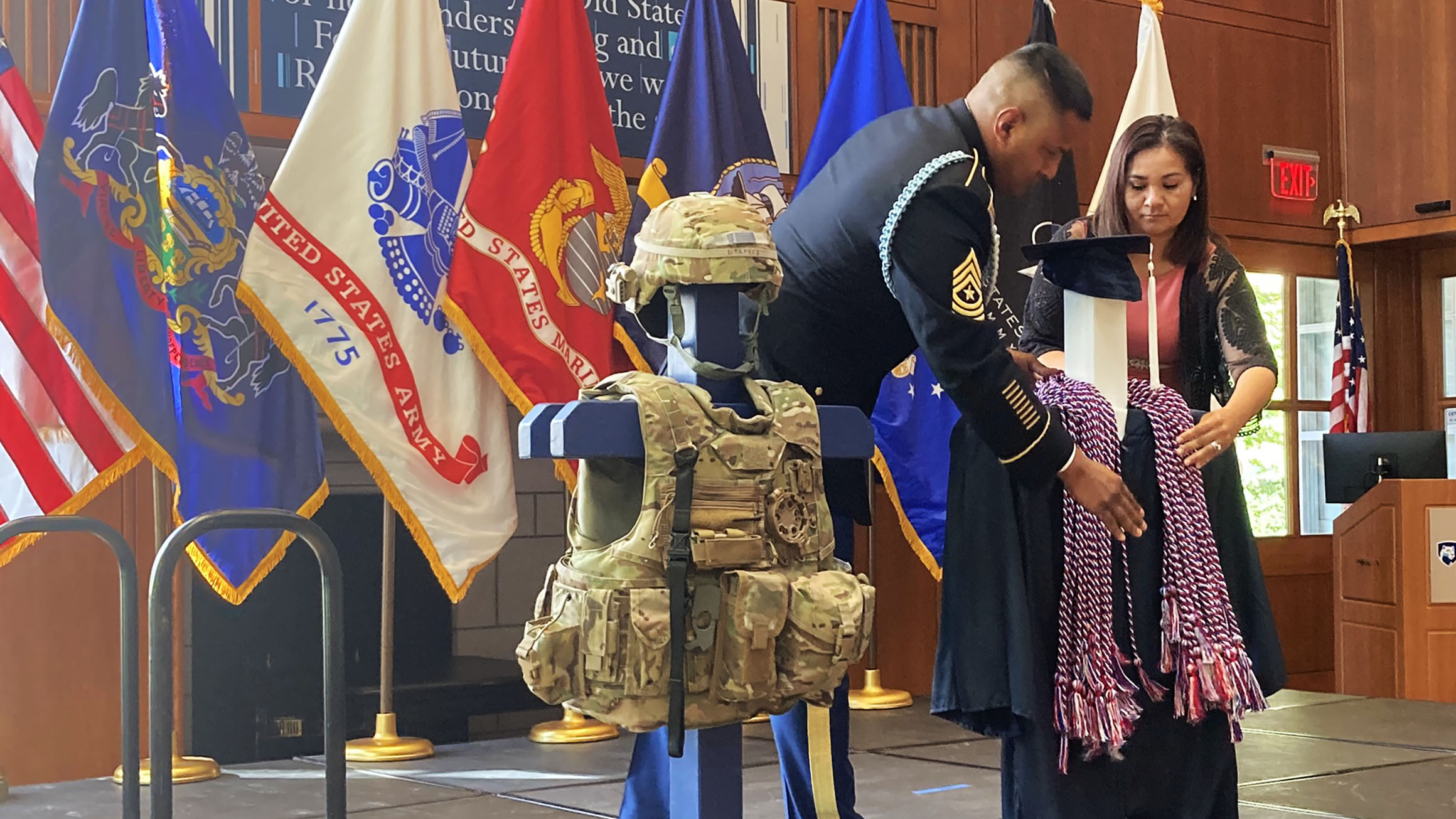 A man and a woman place the honor cords around the neck of a dummy wearing a graduation gown on a stage with flags of the service academies in the background