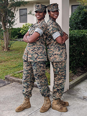 Two women stand back to back wearing military uniforms.
