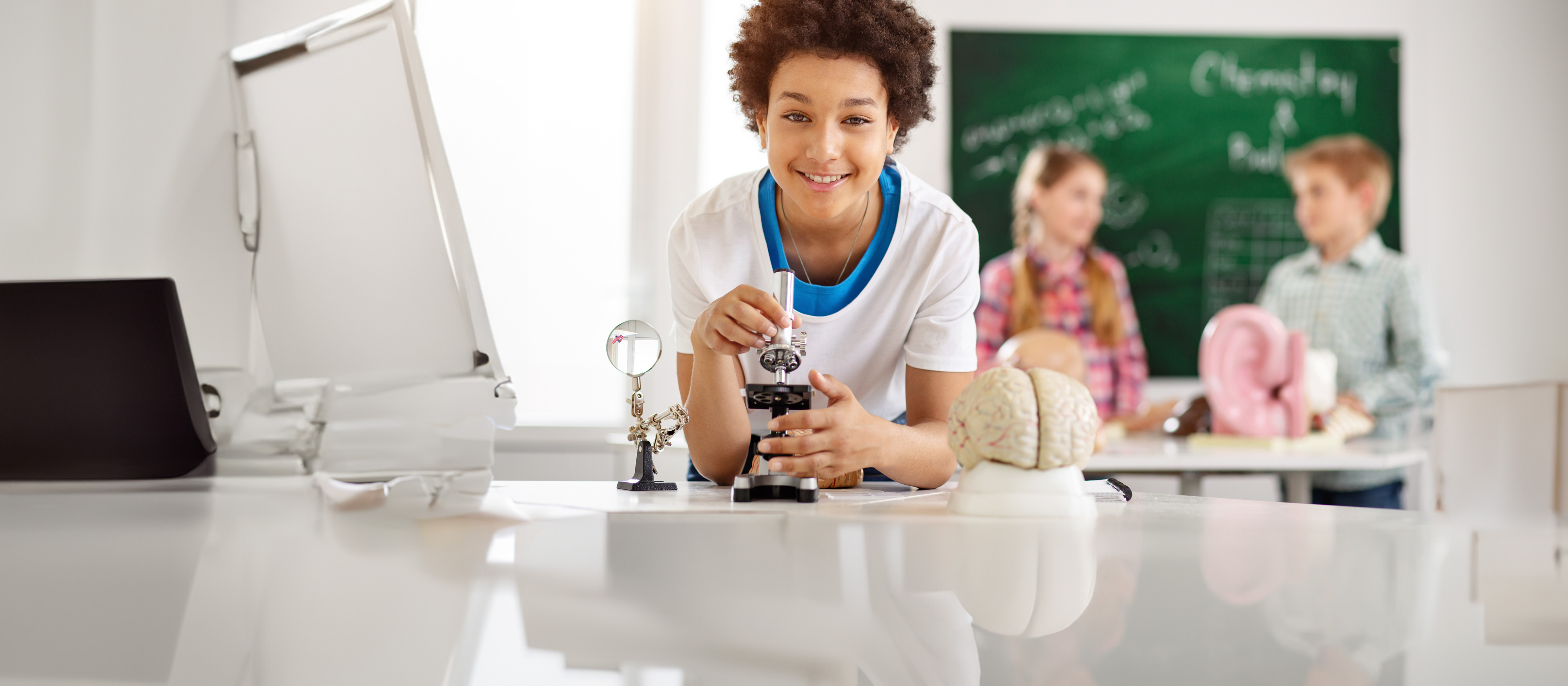 student smiling while holding a microscope in a classroom