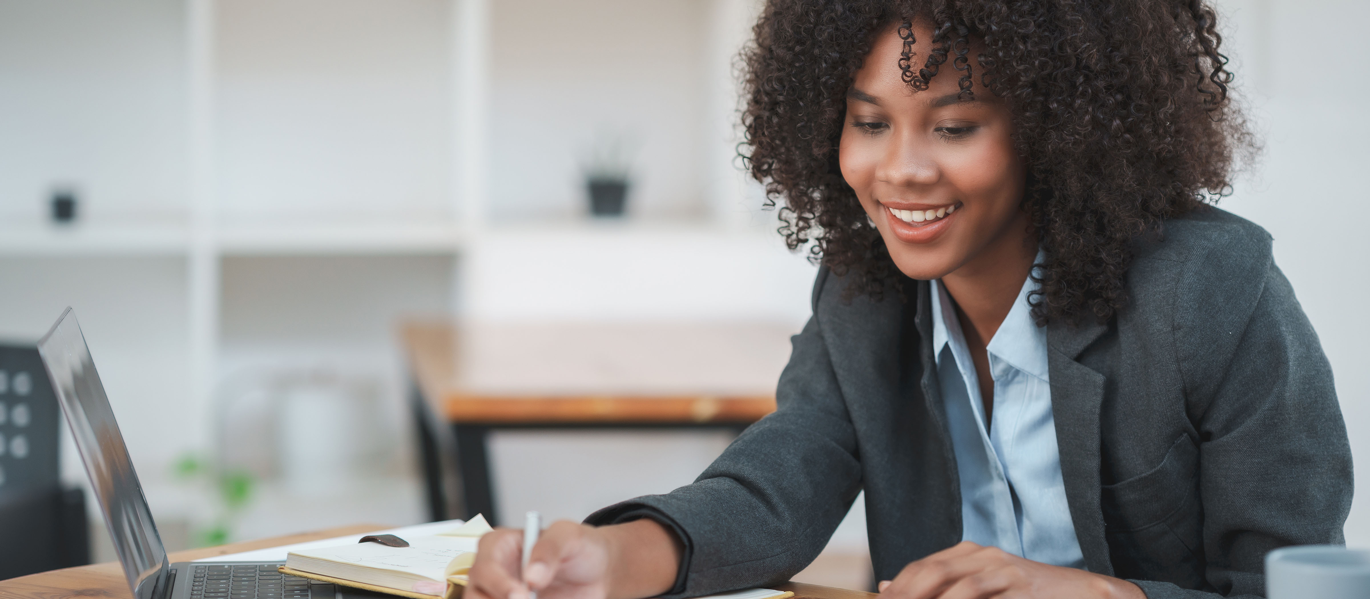 Woman wearing a blazer smiles while writing notes in front of a laptop.