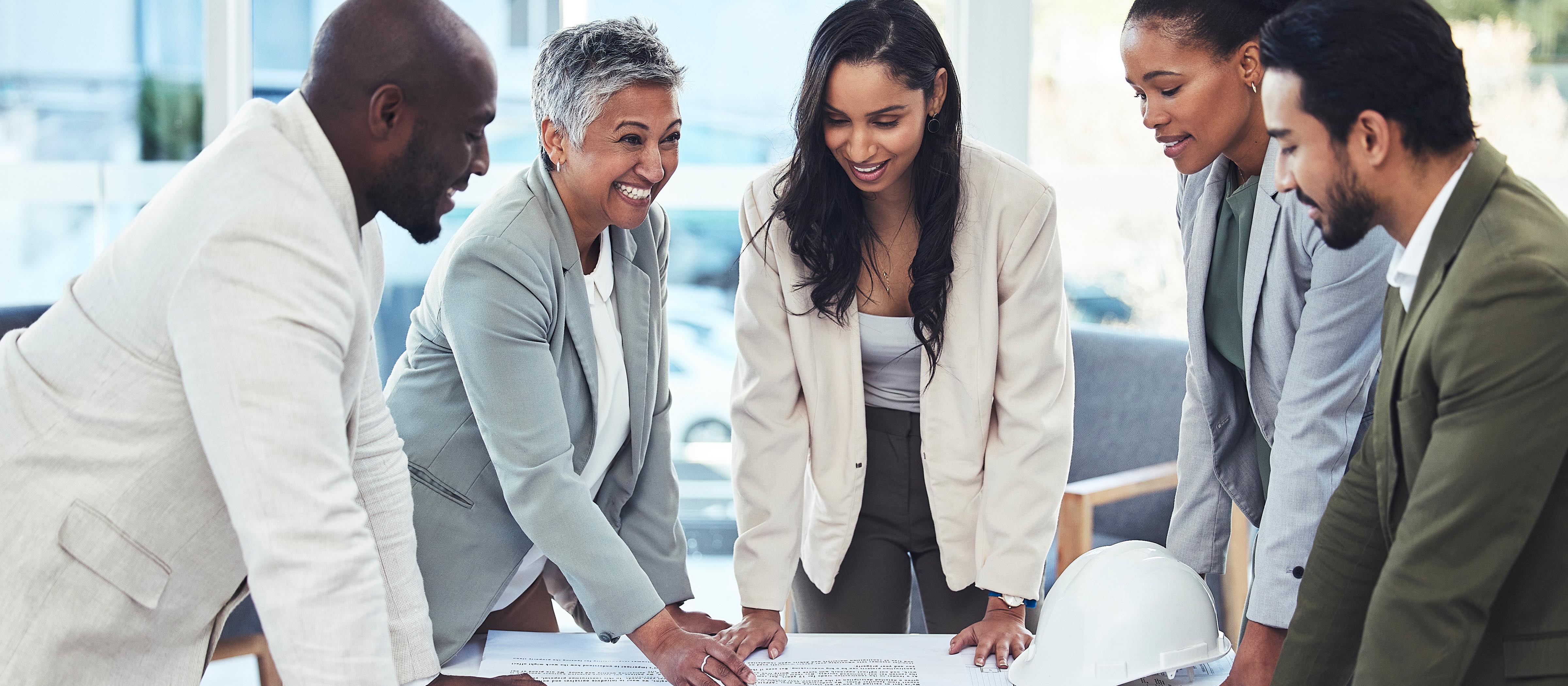 Group of diverse business people stand around documents on a table. One person is smiling at the others.