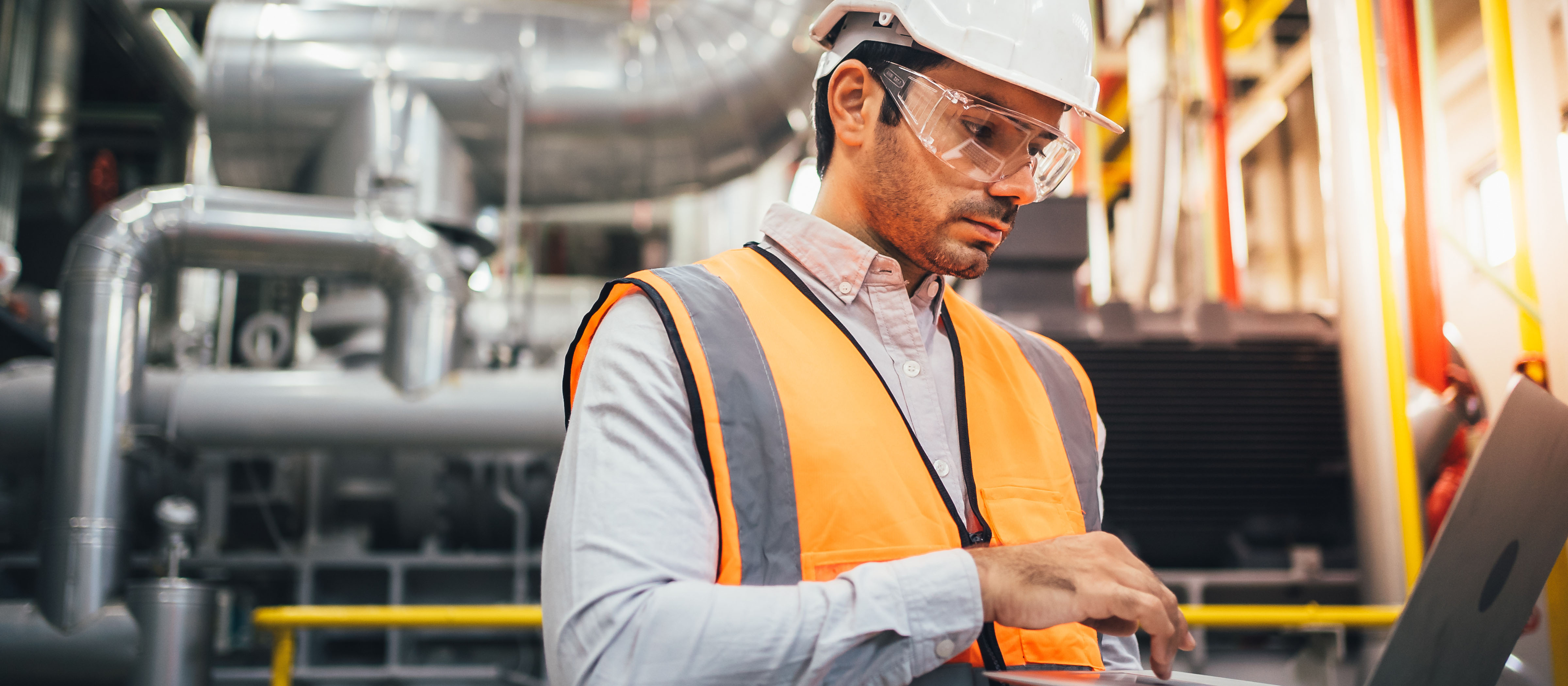 Man in hard hat, an orange construction vest, and a collared shirt works on a laptop while in a factory.