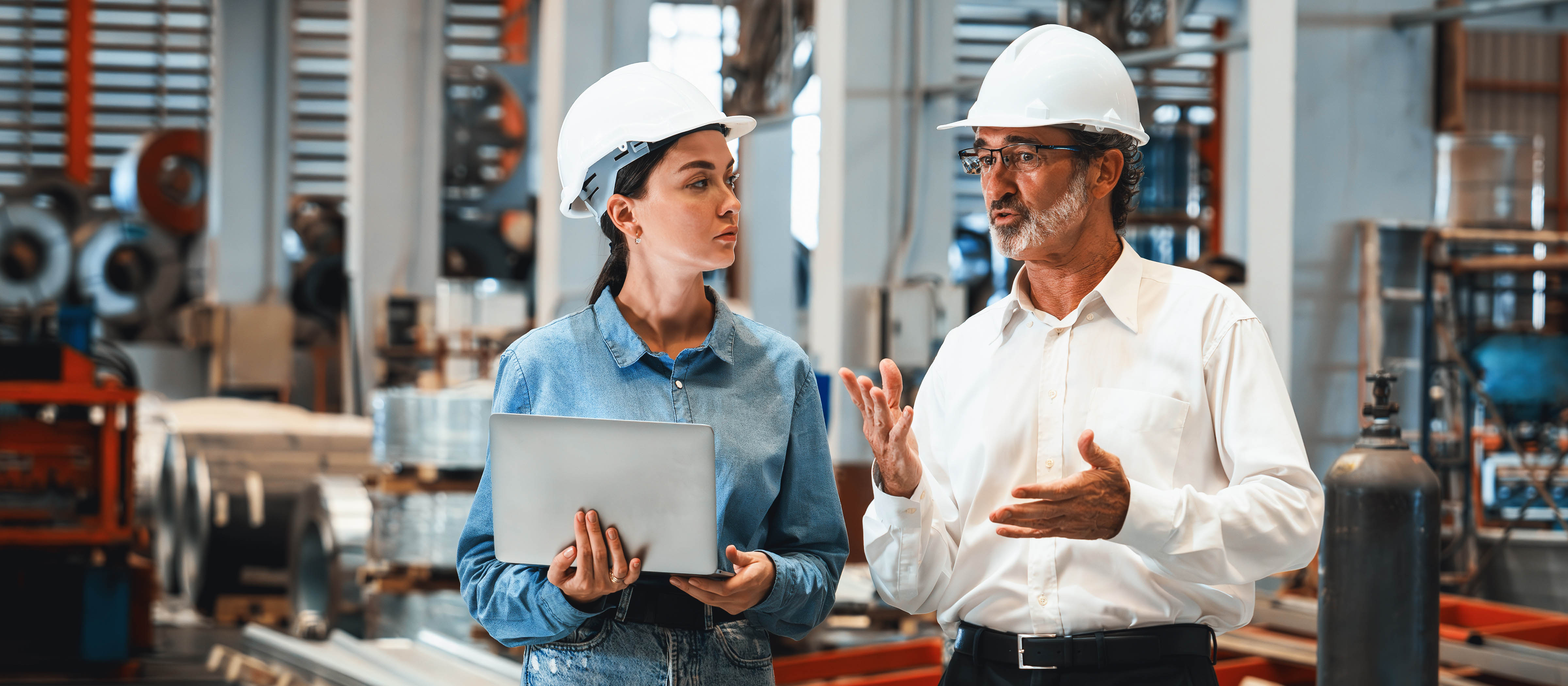 Man and woman in collared shirts and hard hats talk in a factory.