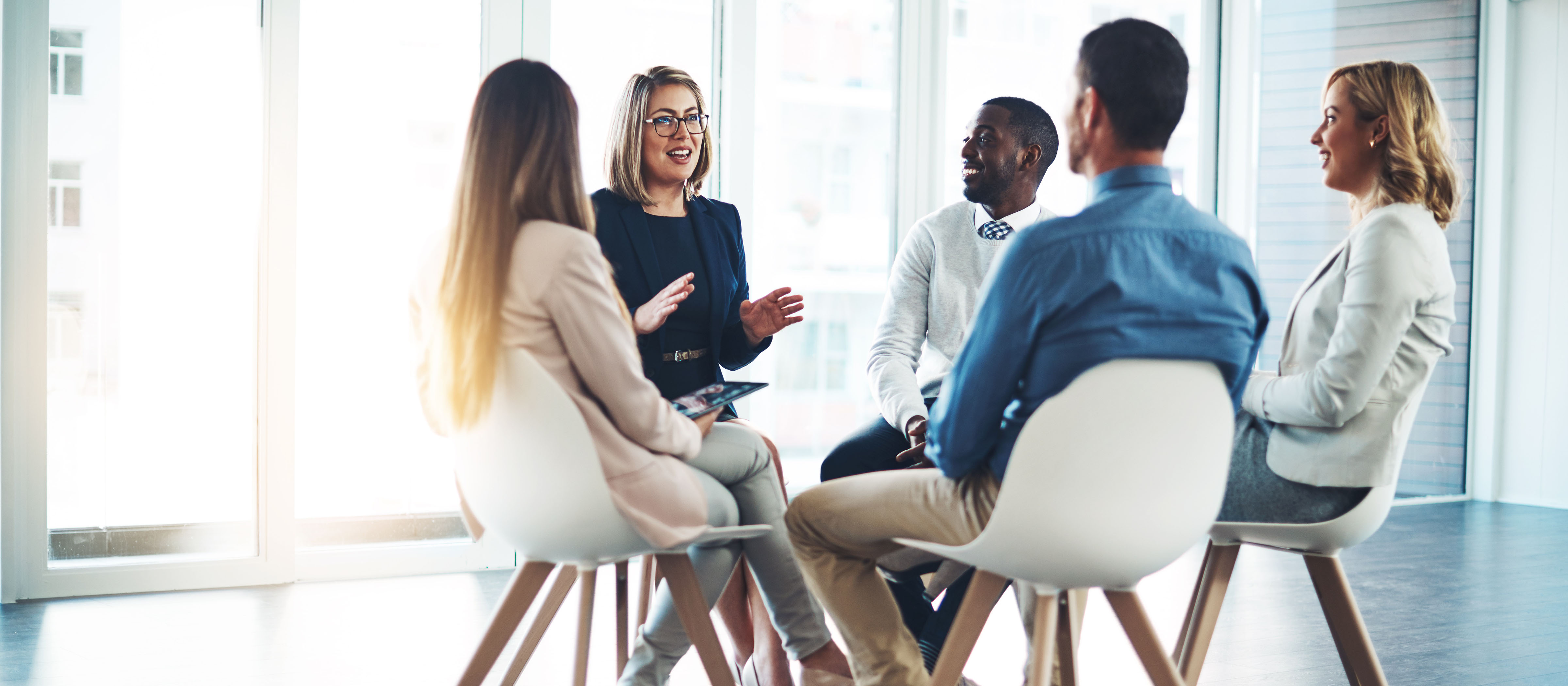 Five people in business casual clothing sit in a tight circle. One woman speaks while the others smile and listen.