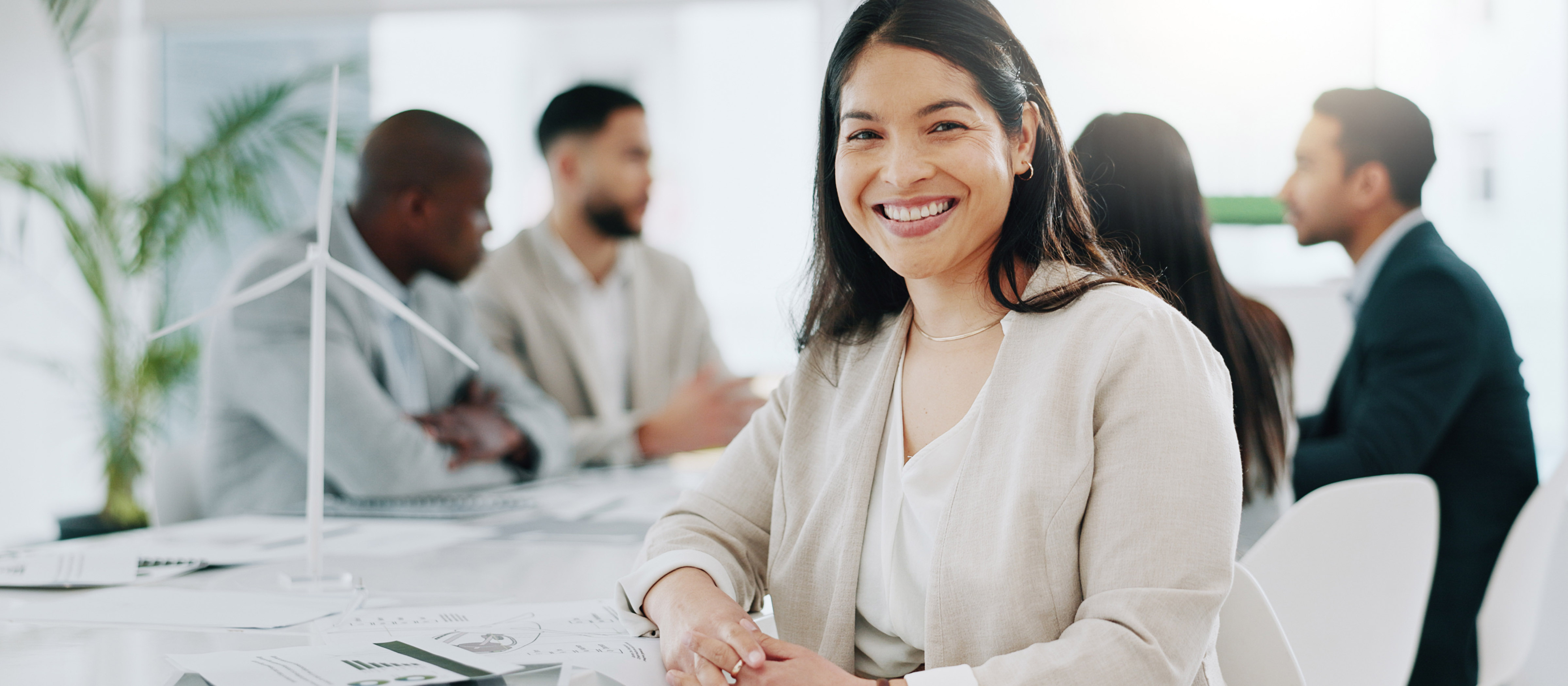 Woman with long dark hair wearing a blazer smiles at the camera. People are sitting at a table and talking behind her.