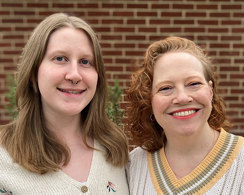 Two women stand in front of a brick wall.