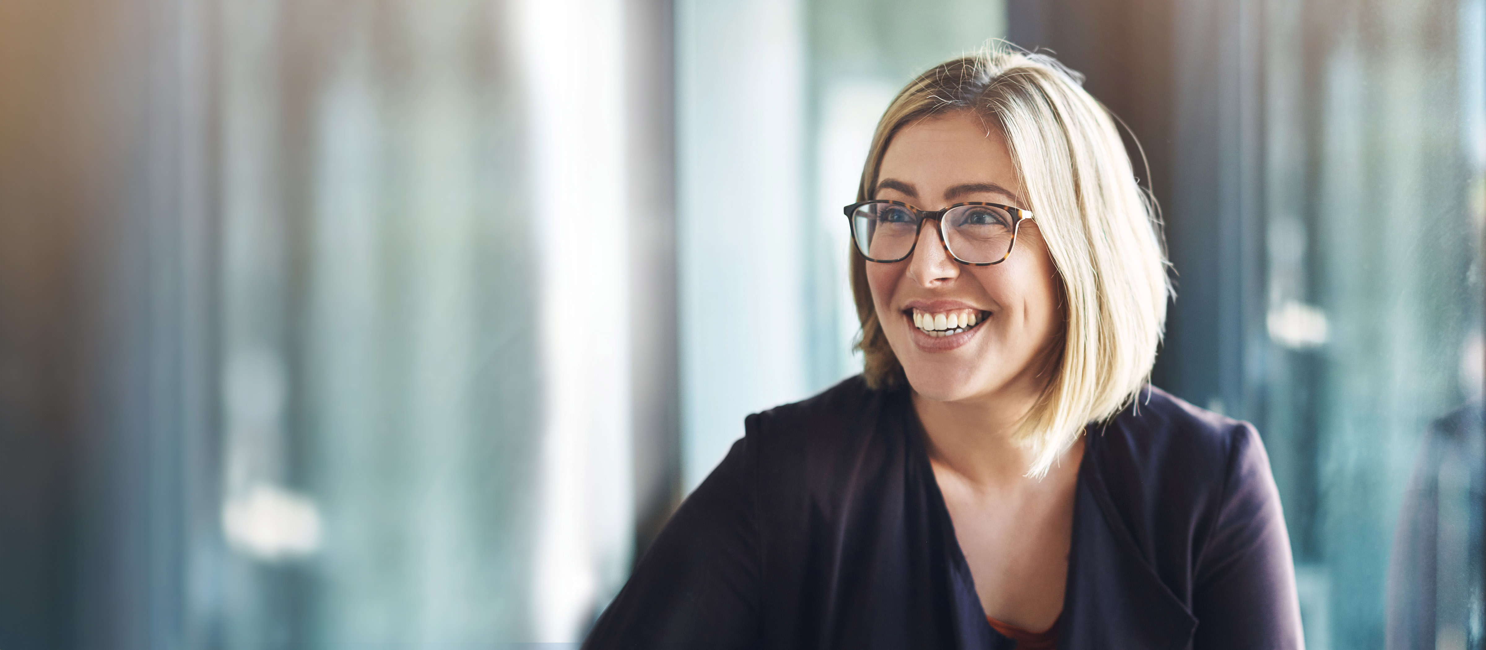 Blond woman wearing glasses smiles at something in the distance. The background behind her is blurry.