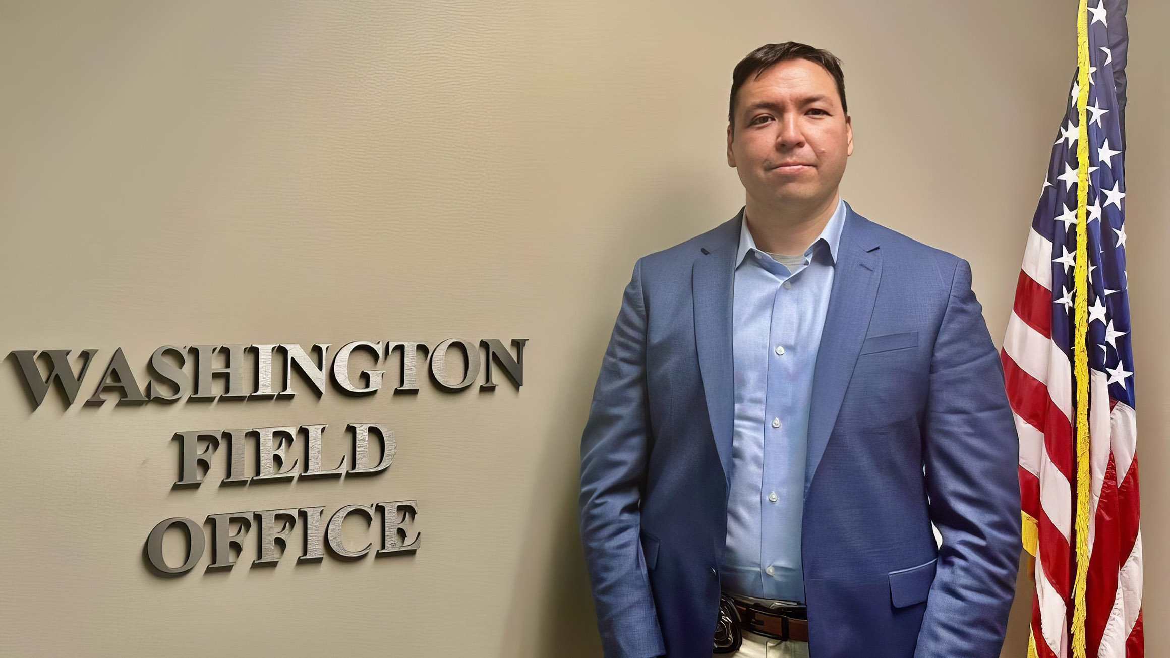 A man in a suit jacket stands against a wall, with a sign that says "Washington Field Office", and a U.S. flag to his right