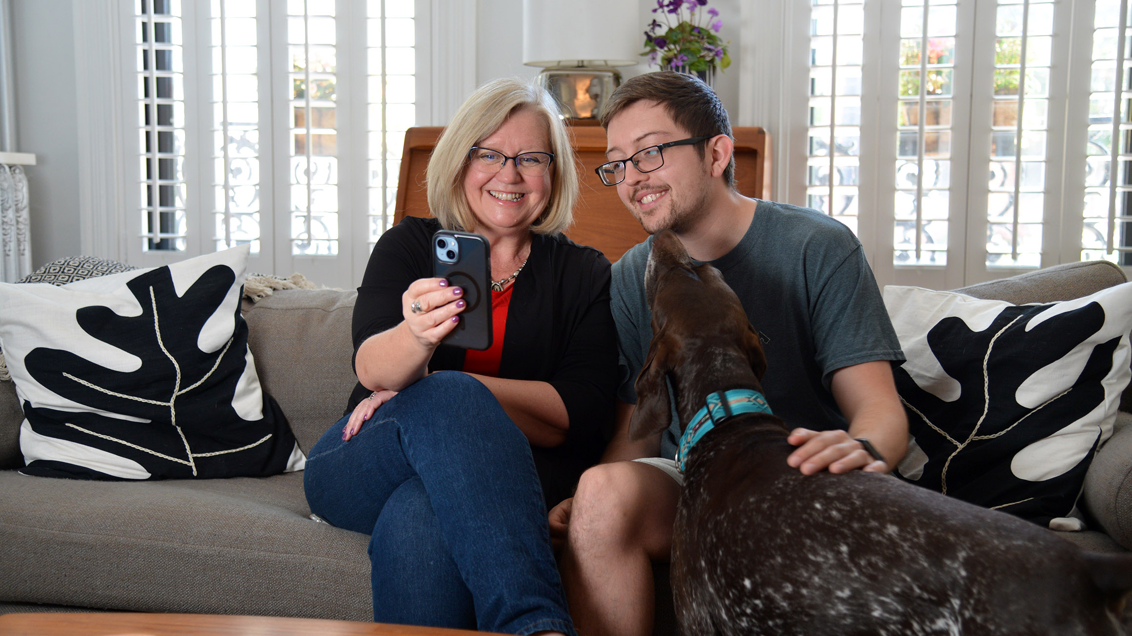 A woman holding a phone sits on a couch with her son as he pets their dog