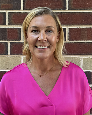 A woman wearing a pink shirt stands in front of a brick wall.