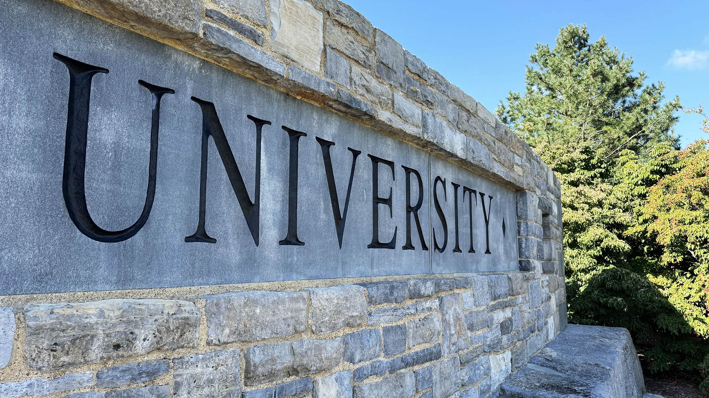 a stone wall shows the word "university" with some trees in the background