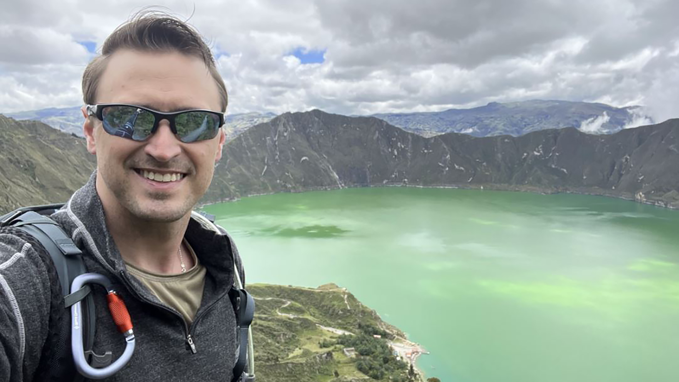 A man takes a selfie in front of a green body of water and mountains.