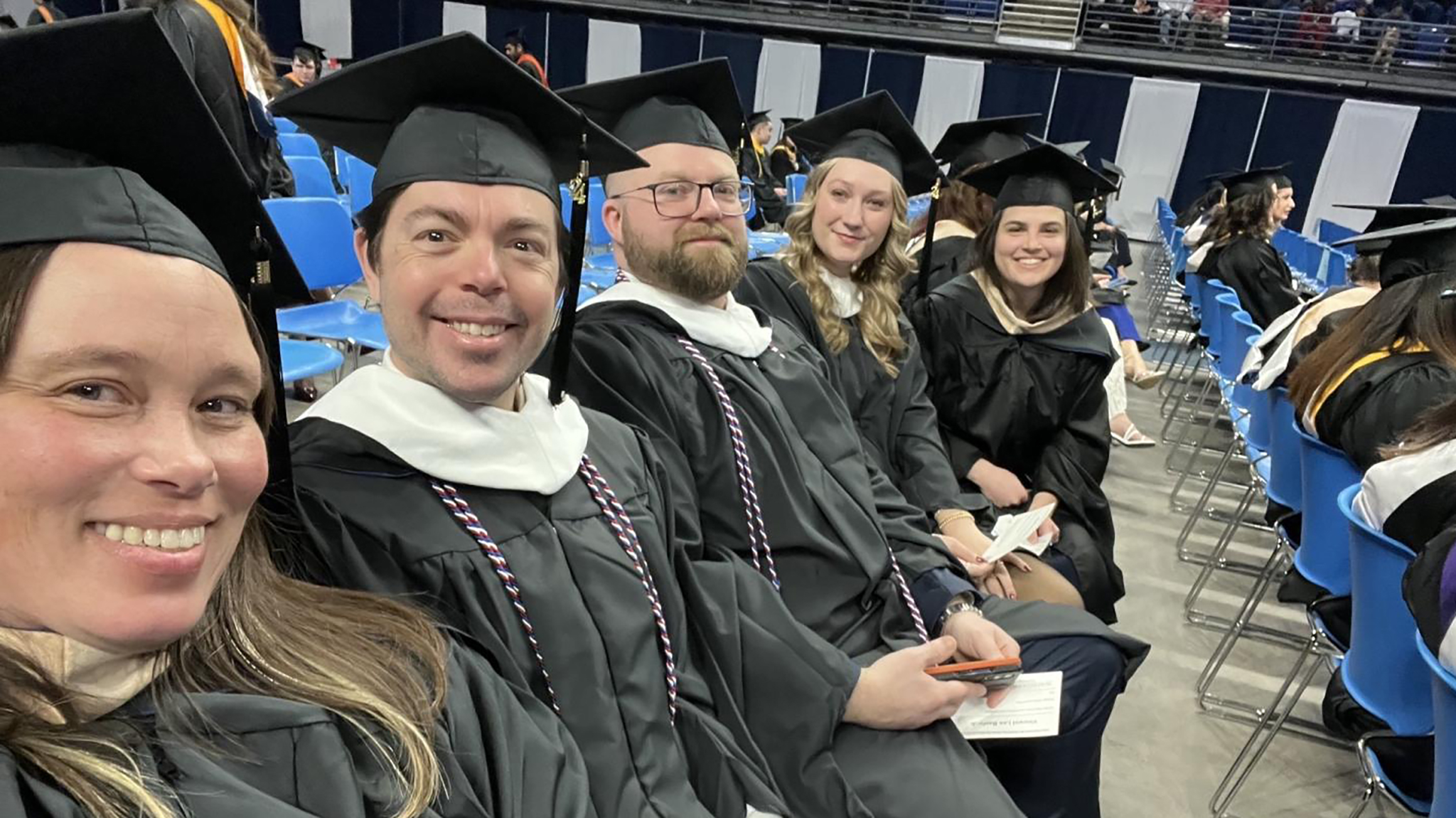 Five people wearing black graduation caps and gowns sit in an arena.