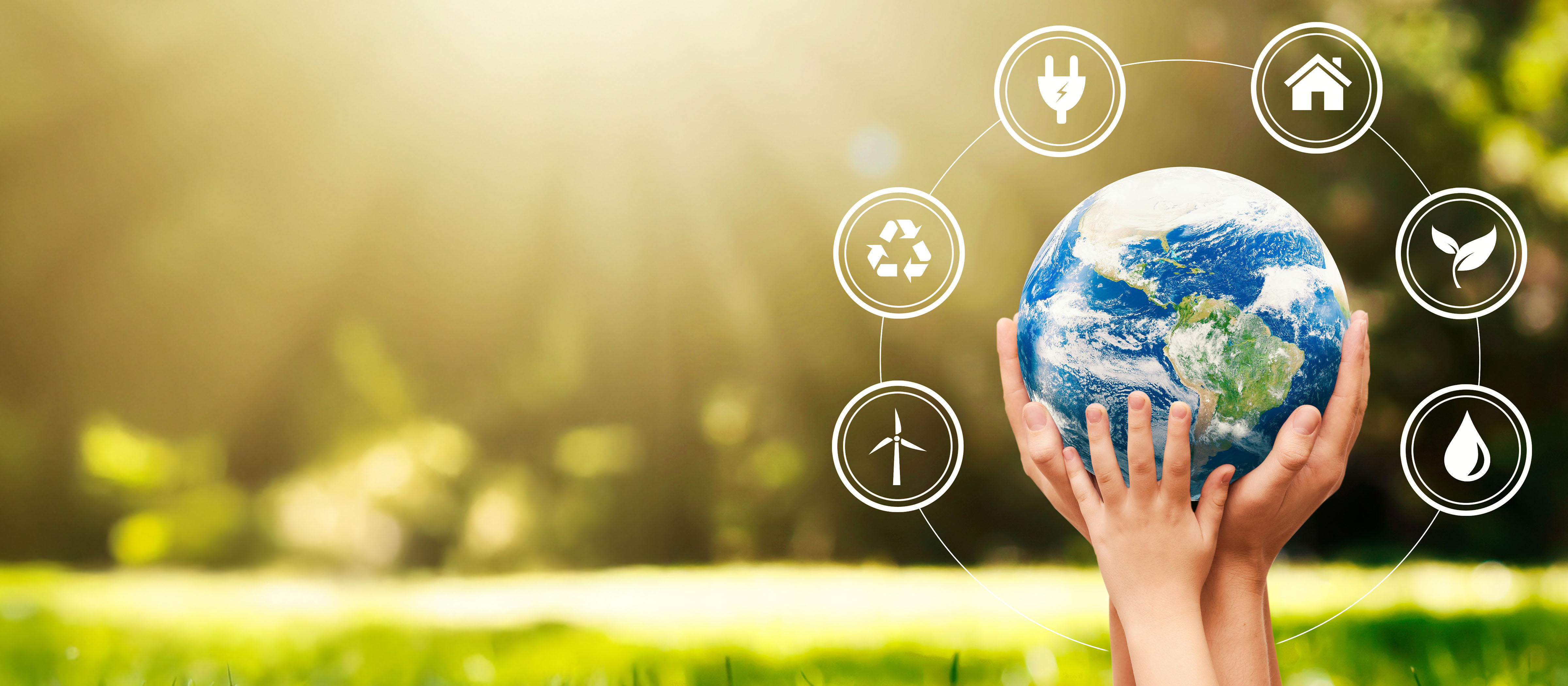 Adult and child hands hold a miniature model of the Earth. Sustainability symbols surround the globe and the sun shine on green grass in the backgorund.