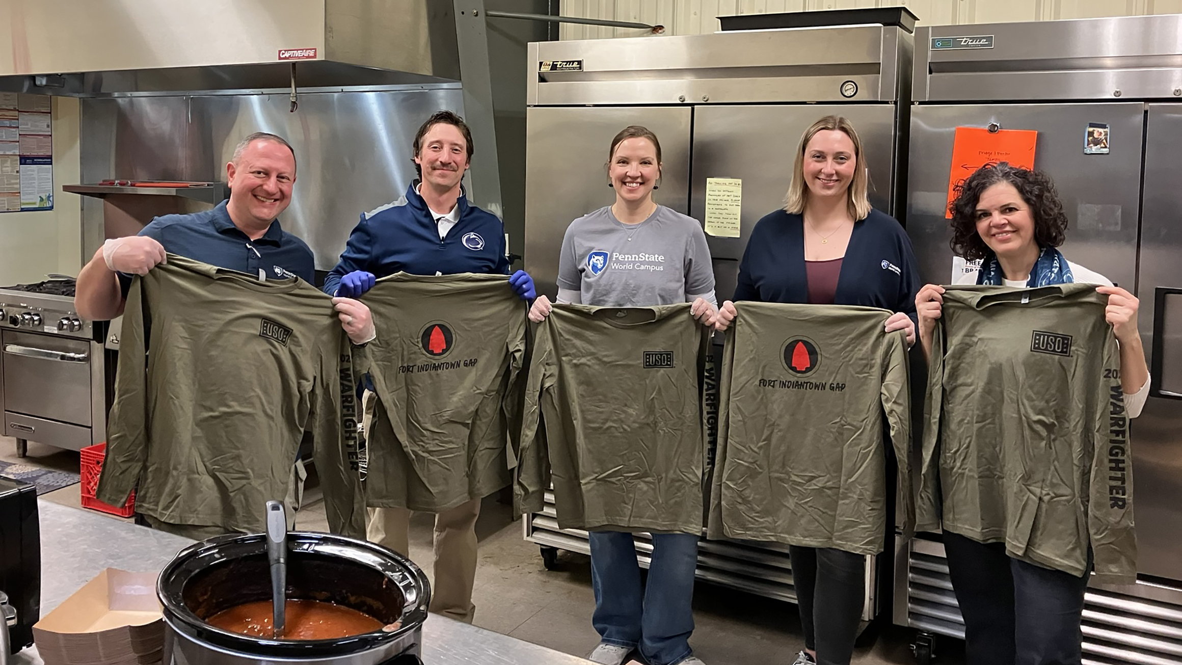 Five people hold long sleeved T-shirts in a kitchen area.
