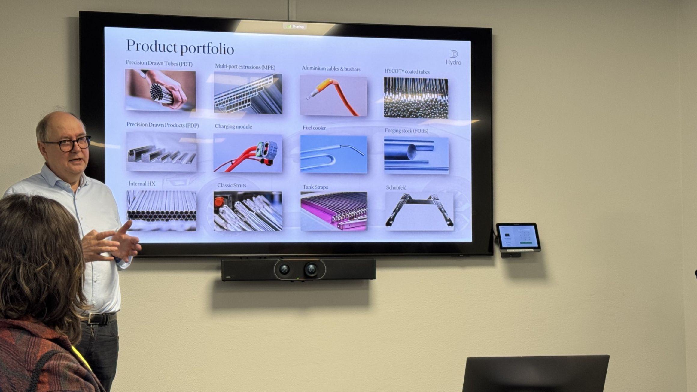 a man in a conference room stands to the left of a monitor showing a presentation about products