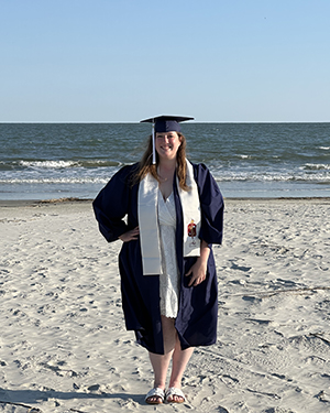 A woman wearing a blue graduation cap and gown on a beach.