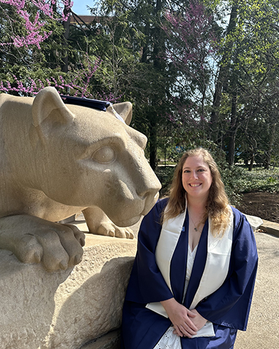 A woman wearing a blue graduation cap and gown sits next to the Nittany Lion Shrine.