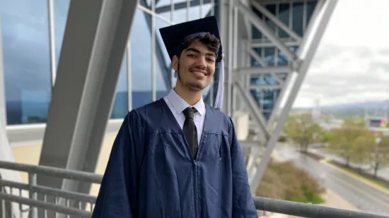 AbdAllah Mahassen, wearing a blue gap and gown, stands along a railing with the exterior of Beaver Stadium in the background