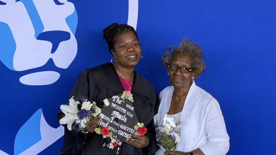 Alicia Douglas, wearing a black gown, poses with her mother. She is holding a graduation cap with an inspirational message that the degree was worth the wait.