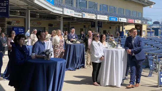 People stand at tables on the concourse of a baseball stadium, listening to a speaker