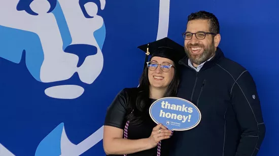 Grace Hussein, wearing a black graduation cap and gown, poses for a photo with her husband. She is holding a sign that says "thanks, honey"