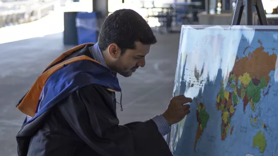 A man in a graduation gown pins a point on a map.