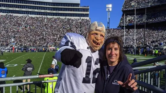 Theresa Tama poses with a fan dressed as a Penn State football player inside Beaver Stadium with the field and stands in the background.