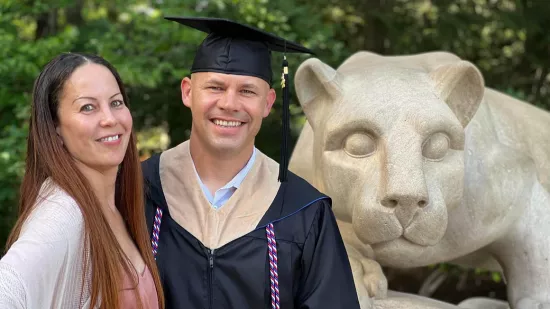 Curt Rice is wearing a cap and gown, posing with his wife, standing in front of the Nittany Lion statue