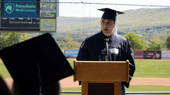 A man in a graduation cap and gown speaks at a podium.