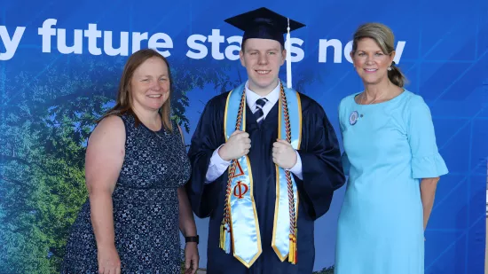 Two women and a man wearing a graduation gown pose in front of a Penn State–themed backdrop.